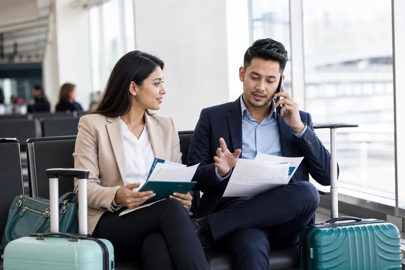Two employees waiting with travel documents and light luggage in an airport lounge